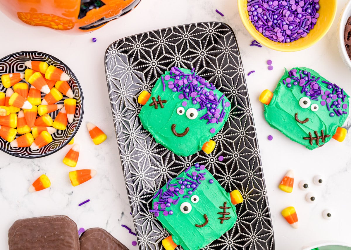 picture of chocolate dipped cookies, covered in green icing with purple sprinkles for hair, candy corn neck bolts, candy eyes, and an icing smile to look like frankenstein