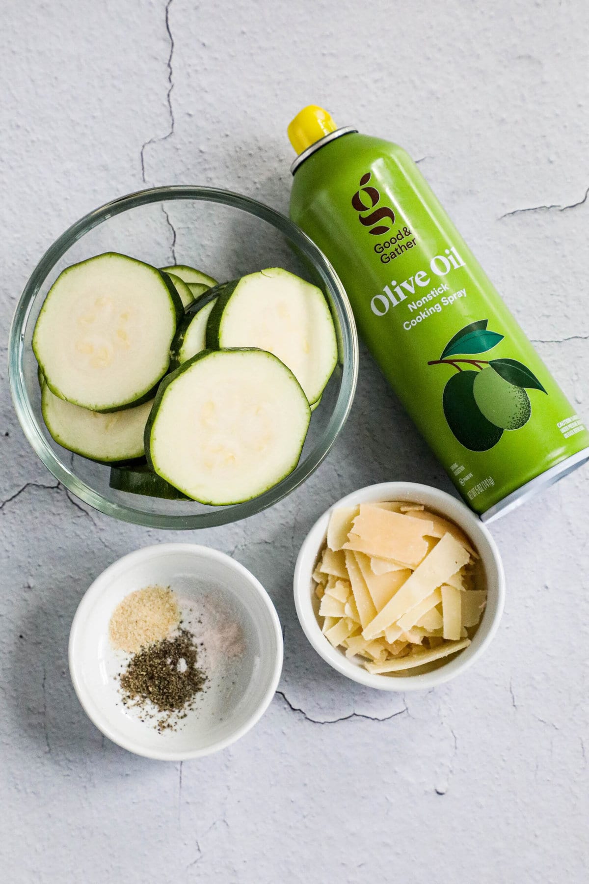 A slice of zucchini next to a bottle of olive oil and other ingredients for a baked parmesan zucchini recipe.