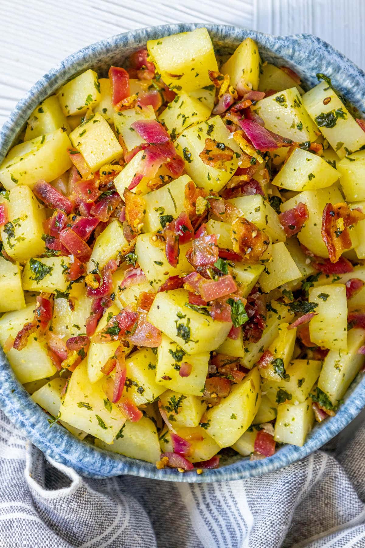 picture of german potato salad with red onions, herbs, and bacon on top in a blue bowl on a table