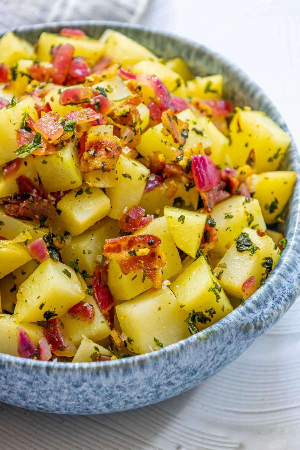 picture of german potato salad with red onions, herbs, and bacon on top in a blue bowl on a table
