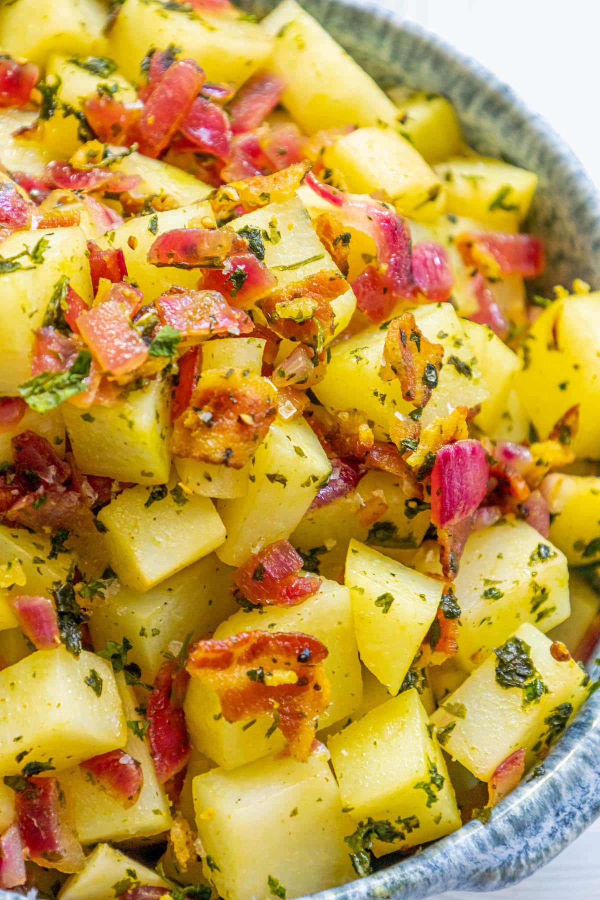 picture of german potato salad with red onions, herbs, and bacon on top in a blue bowl on a table