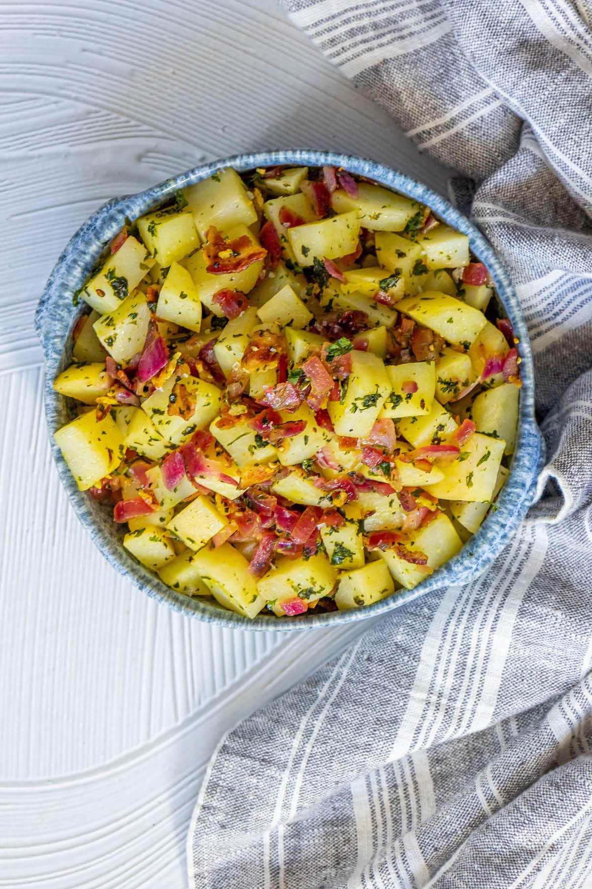 picture of german potato salad with red onions, herbs, and bacon on top in a blue bowl on a table