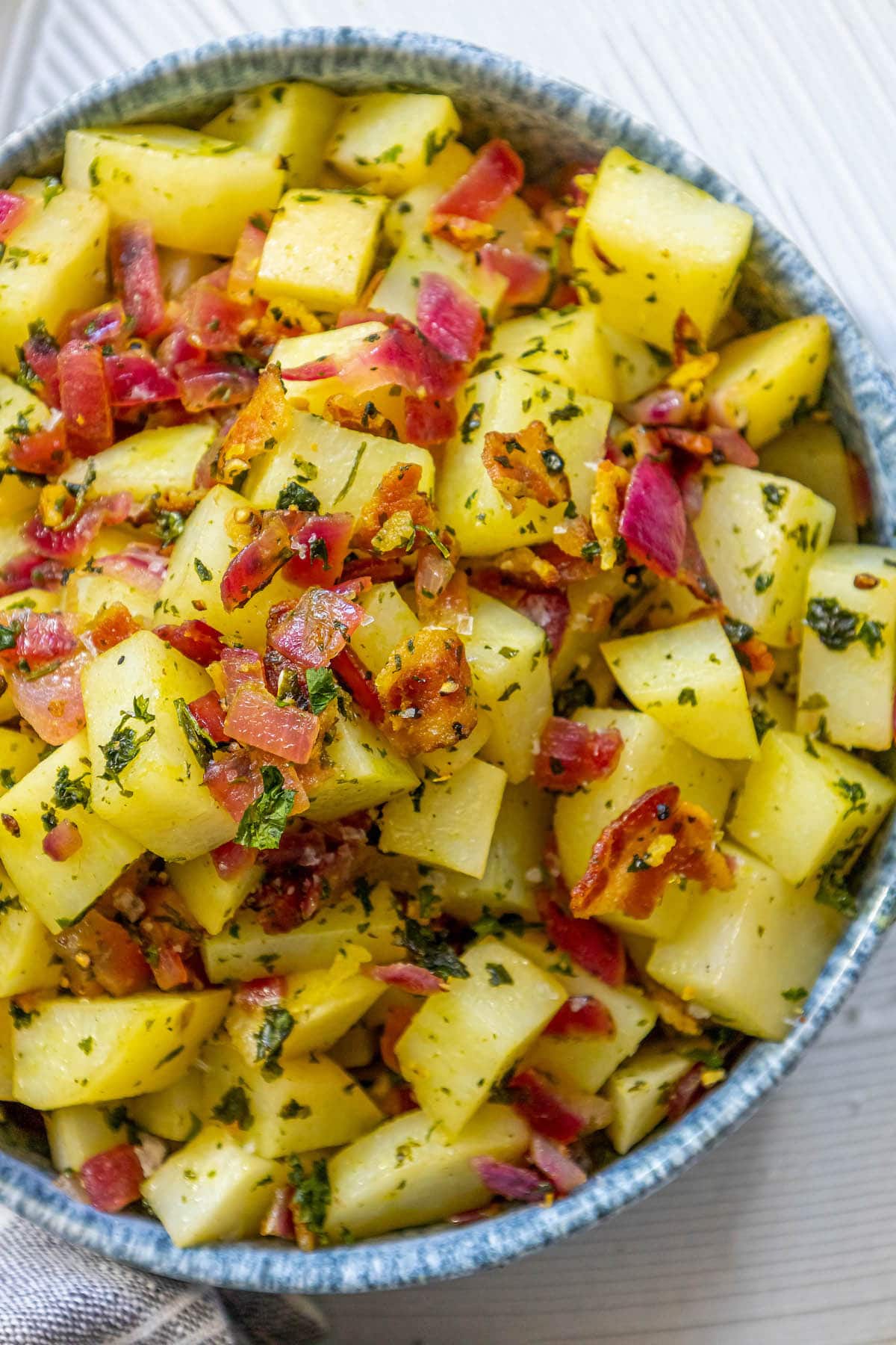 picture of german potato salad with red onions, herbs, and bacon on top in a blue bowl on a table