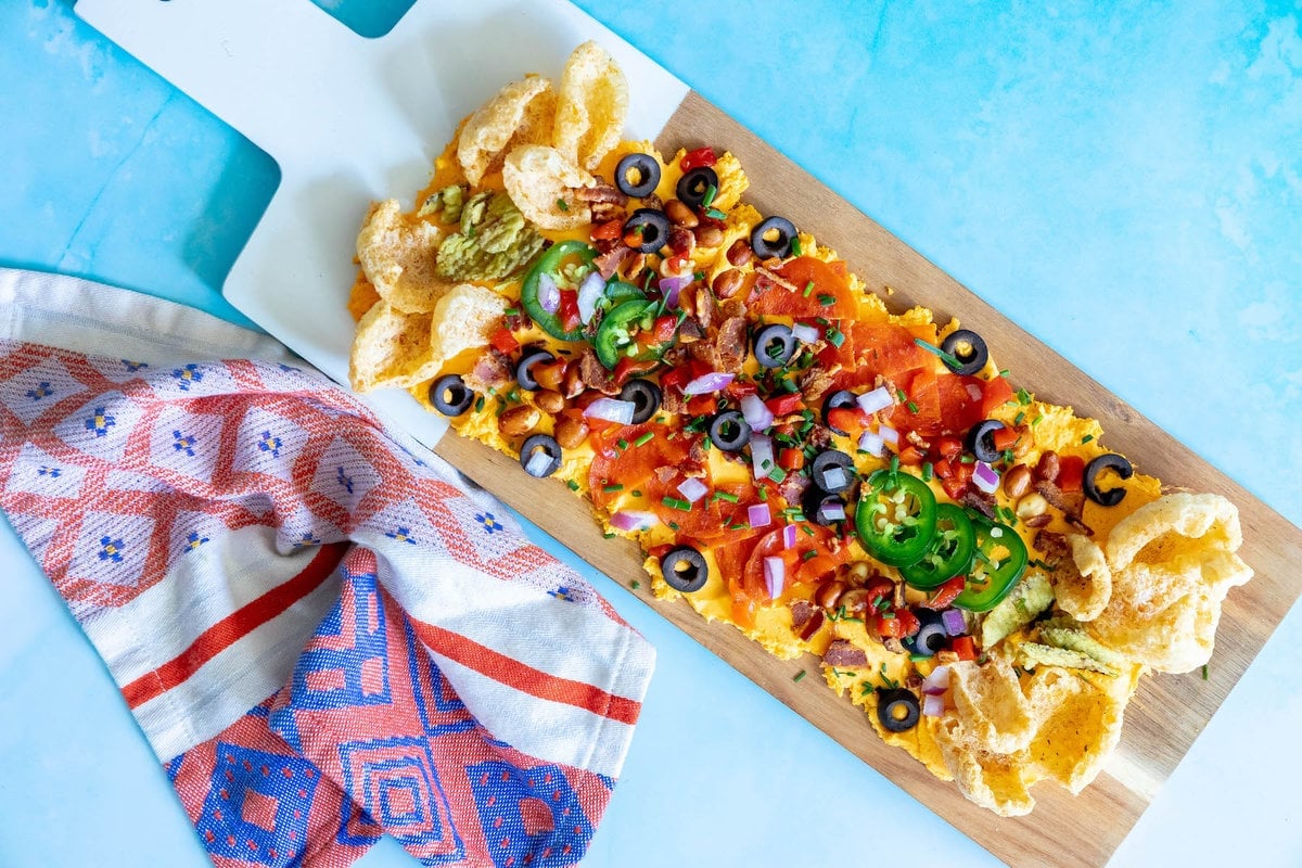 picture of pub cheese spread on a board covered with pork rinds, jalapenos, olives, bacon crumbles, peanuts, pimiento peppers, chives, red onion, and pepperoni