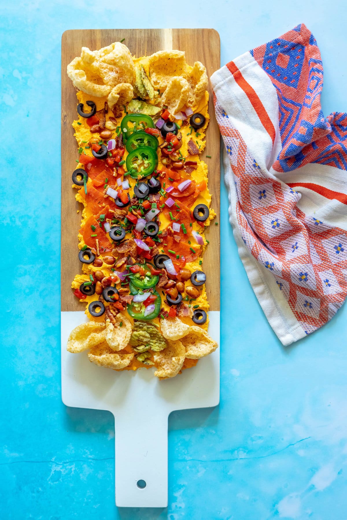 picture of pub cheese spread on a board covered with pork rinds, jalapenos, olives, bacon crumbles, peanuts, pimiento peppers, chives, red onion, and pepperoni