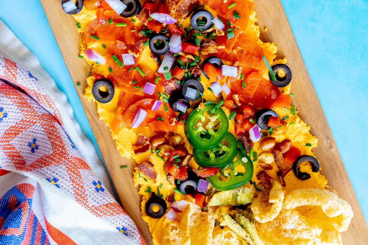 picture of pub cheese spread on a board covered with pork rinds, jalapenos, olives, bacon crumbles, peanuts, pimiento peppers, chives, red onion, and pepperoni