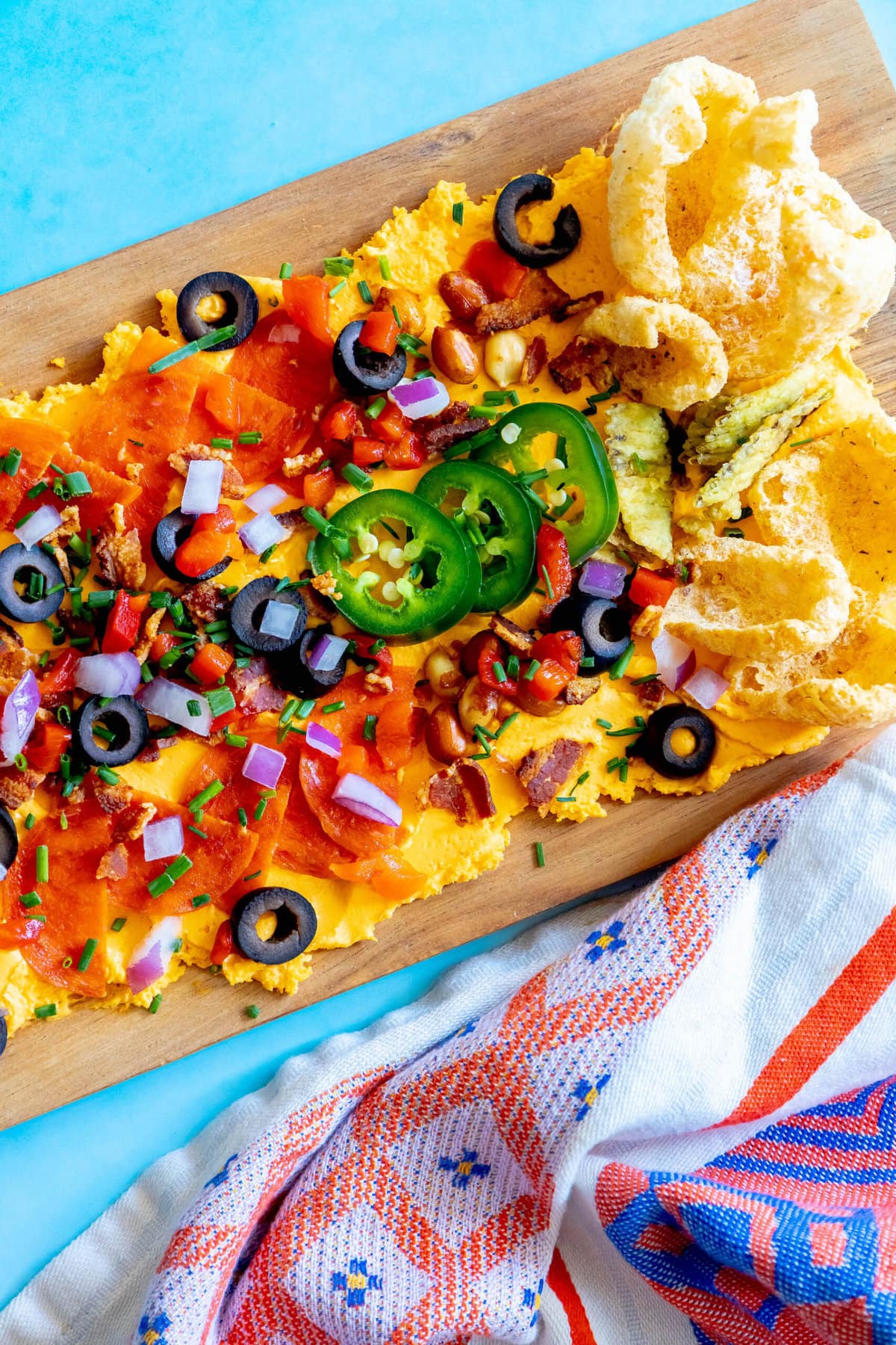 picture of pub cheese spread on a board covered with pork rinds, jalapenos, olives, bacon crumbles, peanuts, pimiento peppers, chives, red onion, and pepperoni