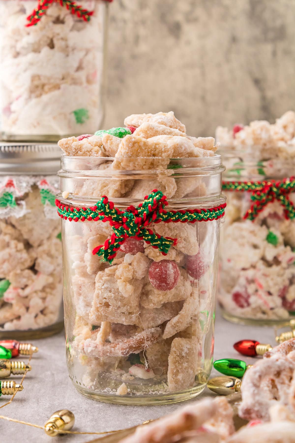 picture of muddy buddies chex mix with pretzels and red and green candies mixed in coated in powdered sugar in a jar with a red and green bow on it 