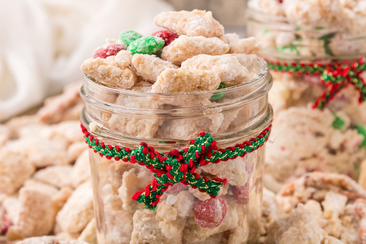 picture of muddy buddies chex mix with pretzels and red and green candies mixed in coated in powdered sugar in a jar with a red and green ribbon around it