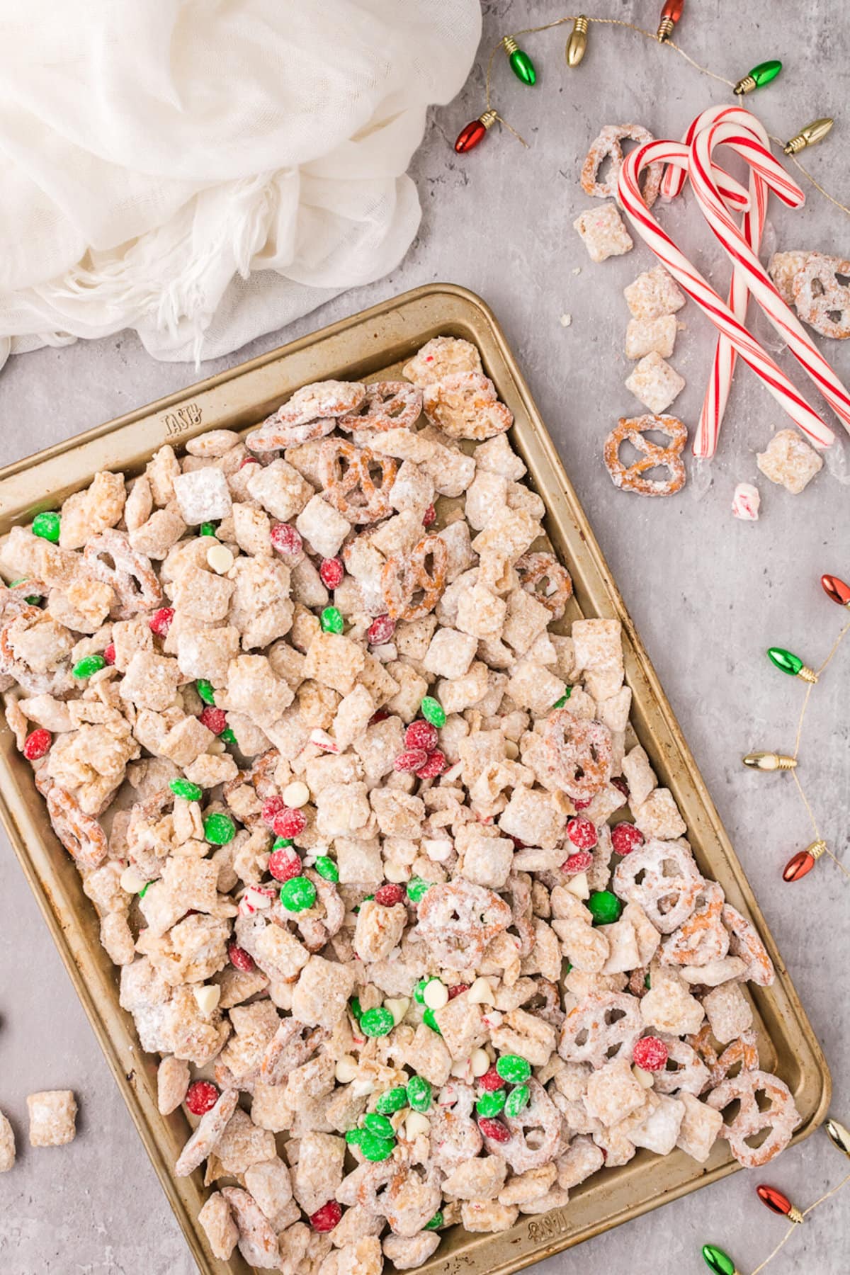 picture of muddy buddies chex mix with pretzels and red and green candies mixed in coated in powdered sugar on a baking sheet