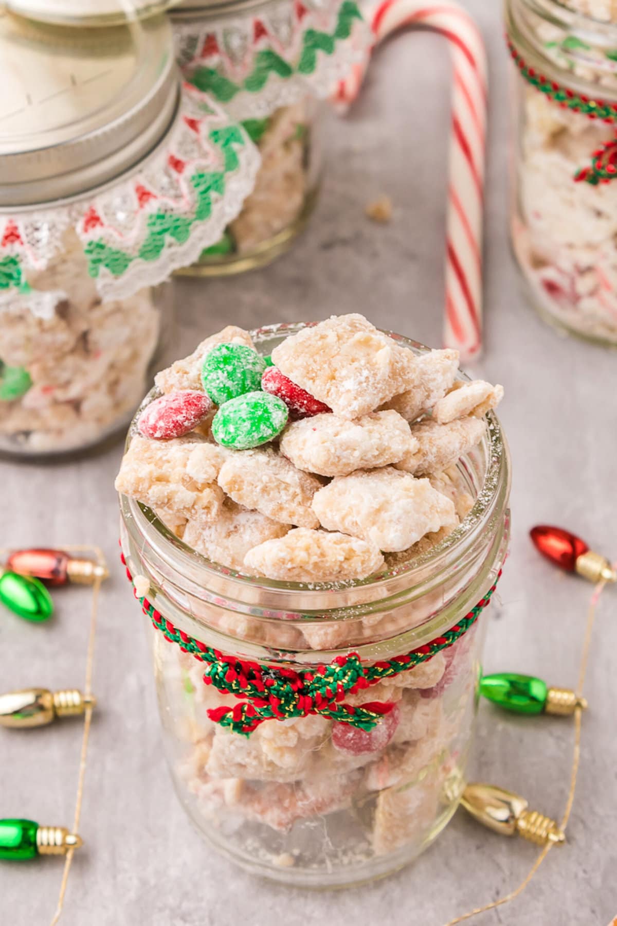 picture of muddy buddies chex mix with pretzels and red and green candies mixed in coated in powdered sugar in a jar with a red and green bow on it 