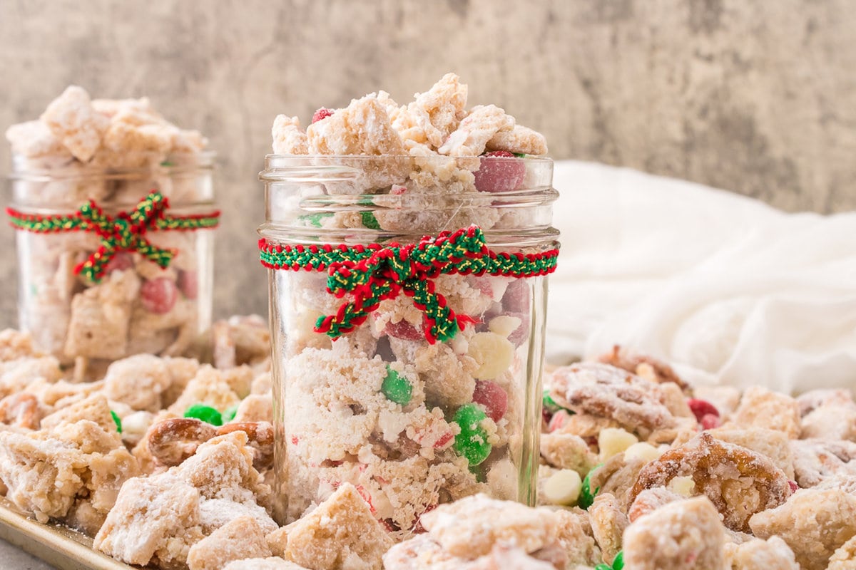 picture of muddy buddies chex mix with pretzels and red and green candies mixed in coated in powdered sugar in a jar with a red and green bow on it 