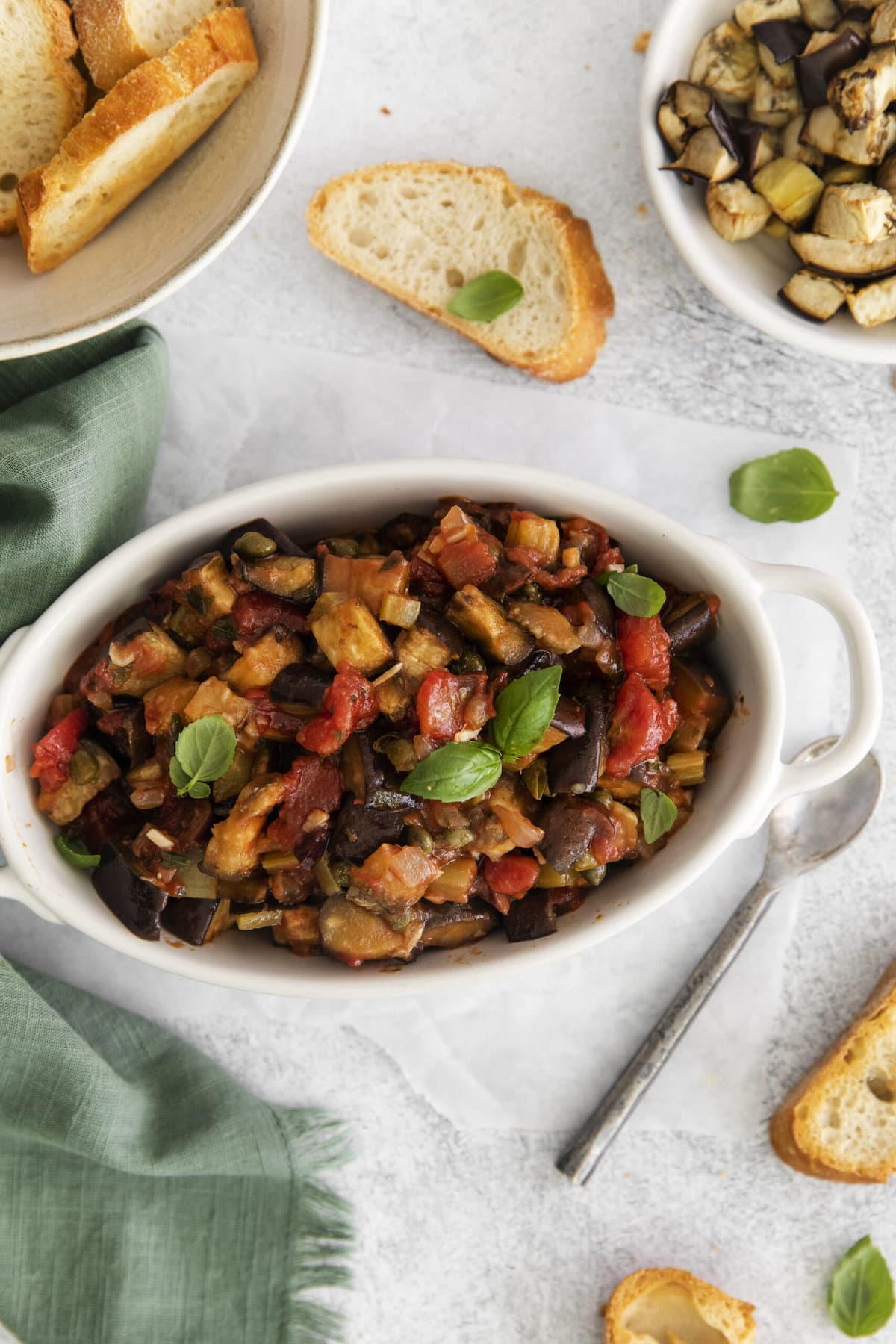 picture of baked eggplant caponata in a white dish with basil leaves on top on a table next to a spoon