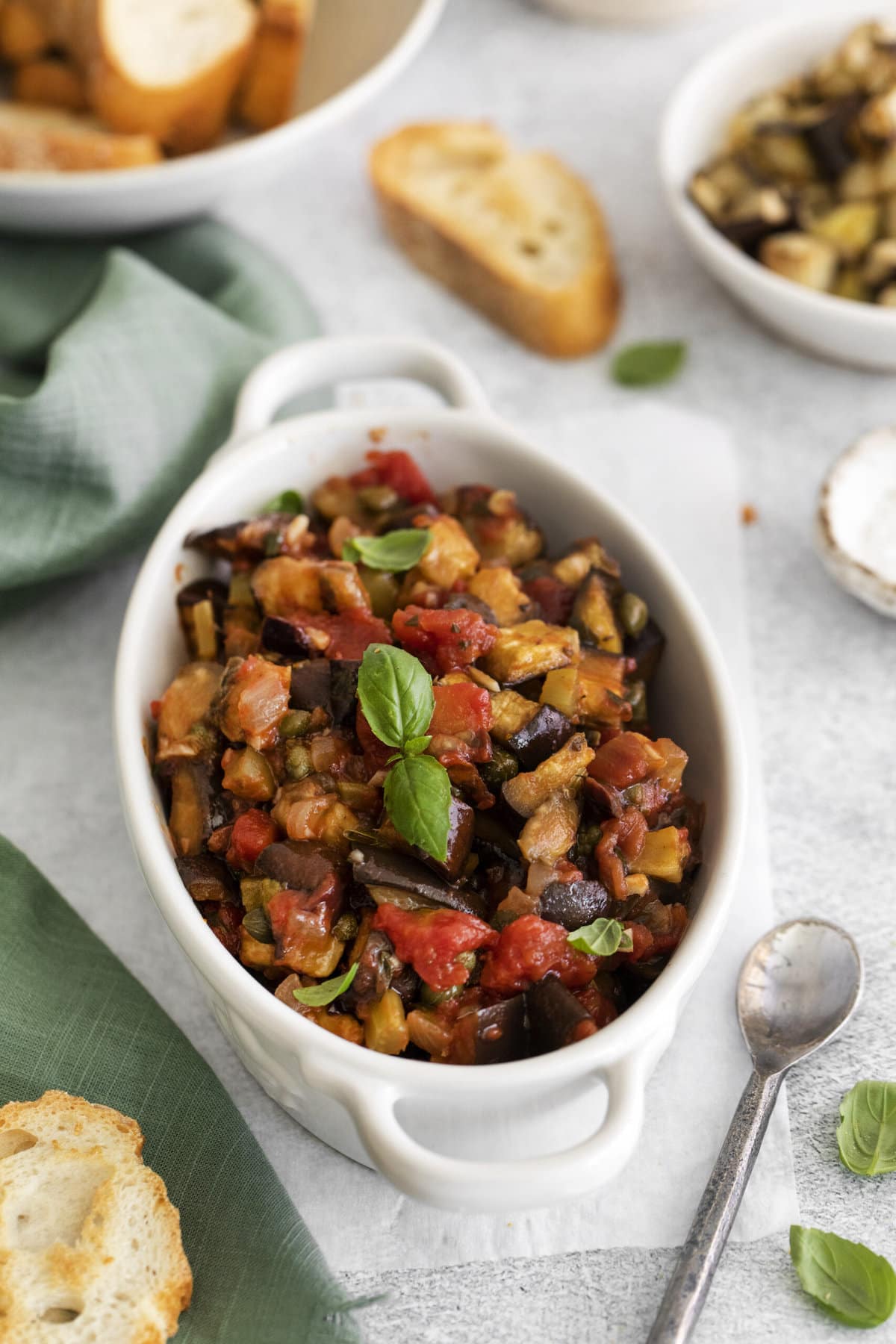 picture of baked eggplant caponata in a white dish with basil leaves on top on a table next to a spoon