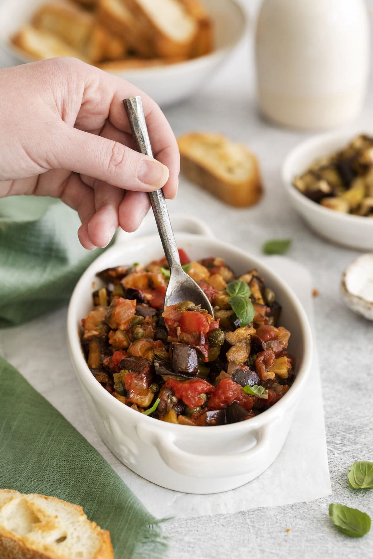 picture of baked eggplant caponata in a white dish with basil leaves on top on a table next to a spoon