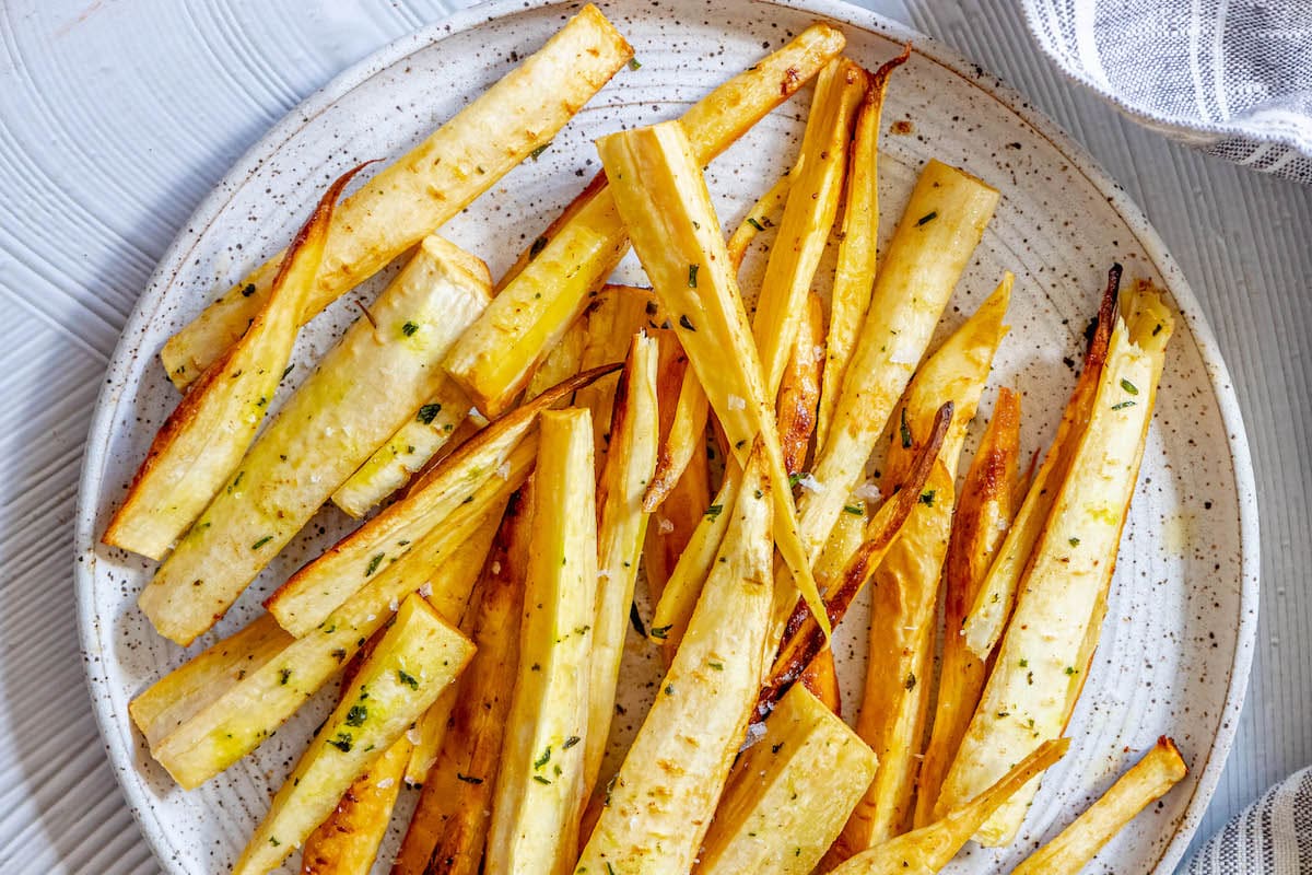 roasted parsnips seasoned with garlic butter and herbs on a white speckled plate next to a blue striped tea towel