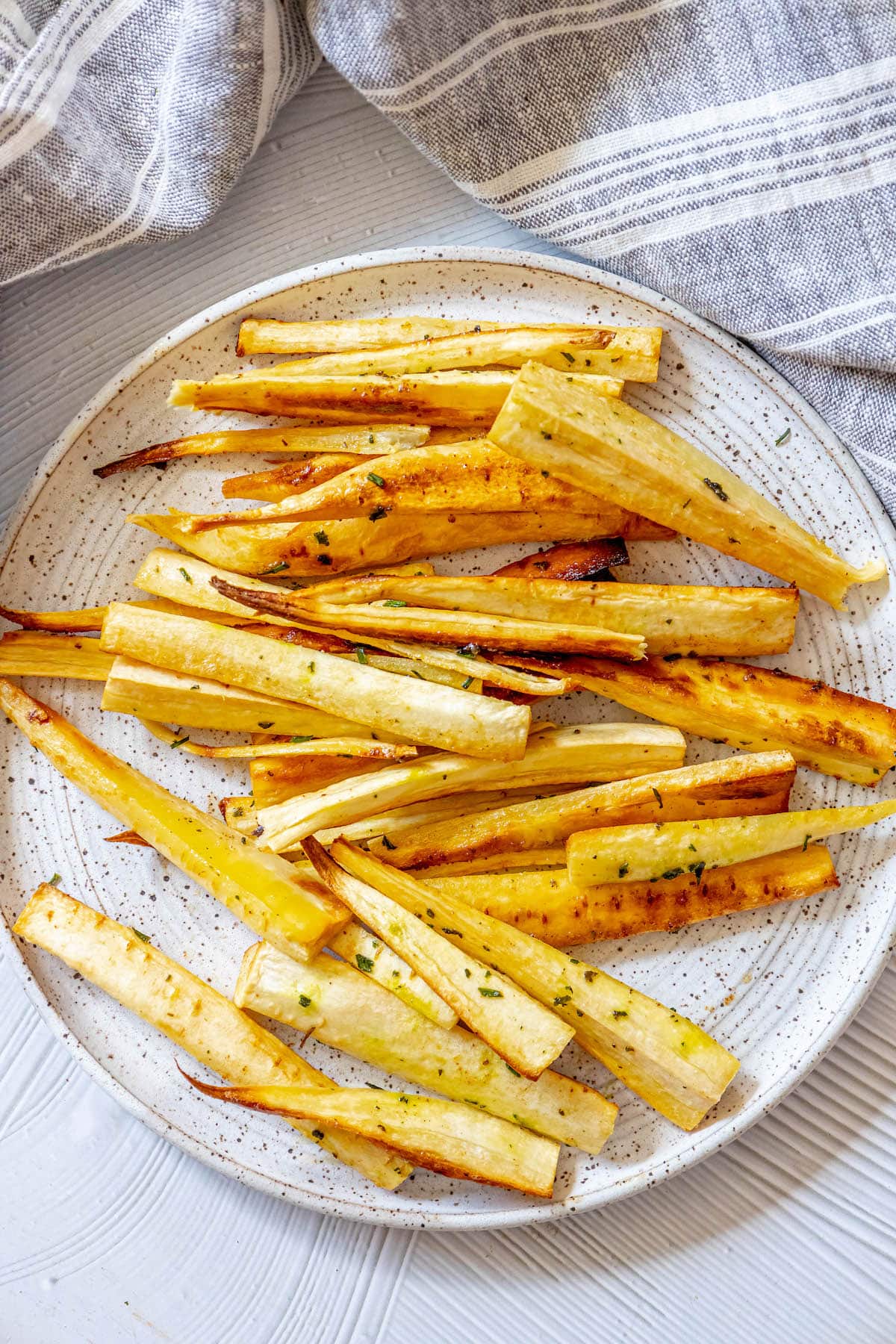 roasted parsnips seasoned with garlic butter and herbs on a white speckled plate next to a blue striped tea towel