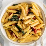 picture of penne pasta in a bowl with kale, sundried tomatoes, and cheese on a table with forks