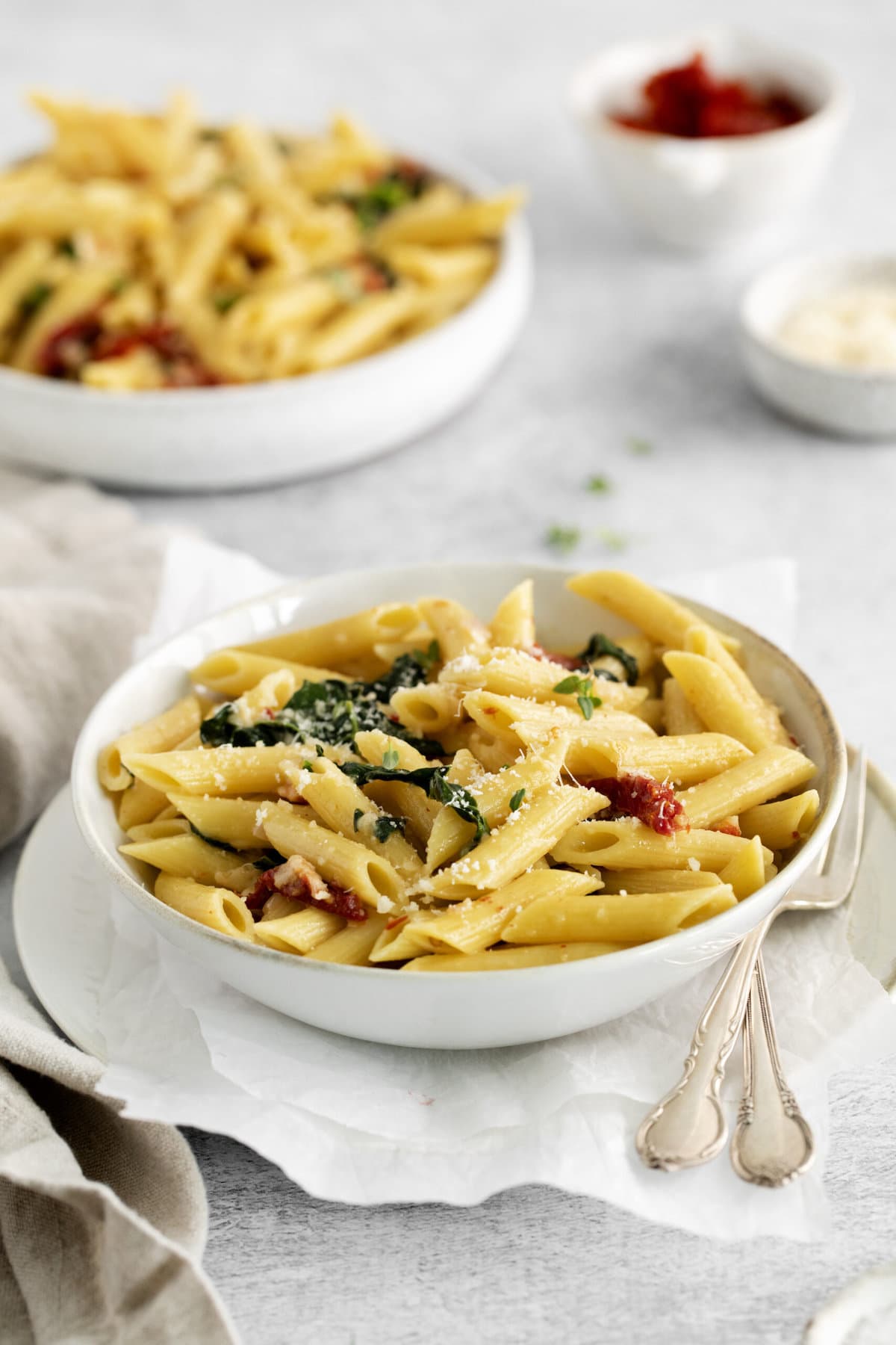 picture of penne pasta in a bowl with kale, sundried tomatoes, and cheese on a table with forks