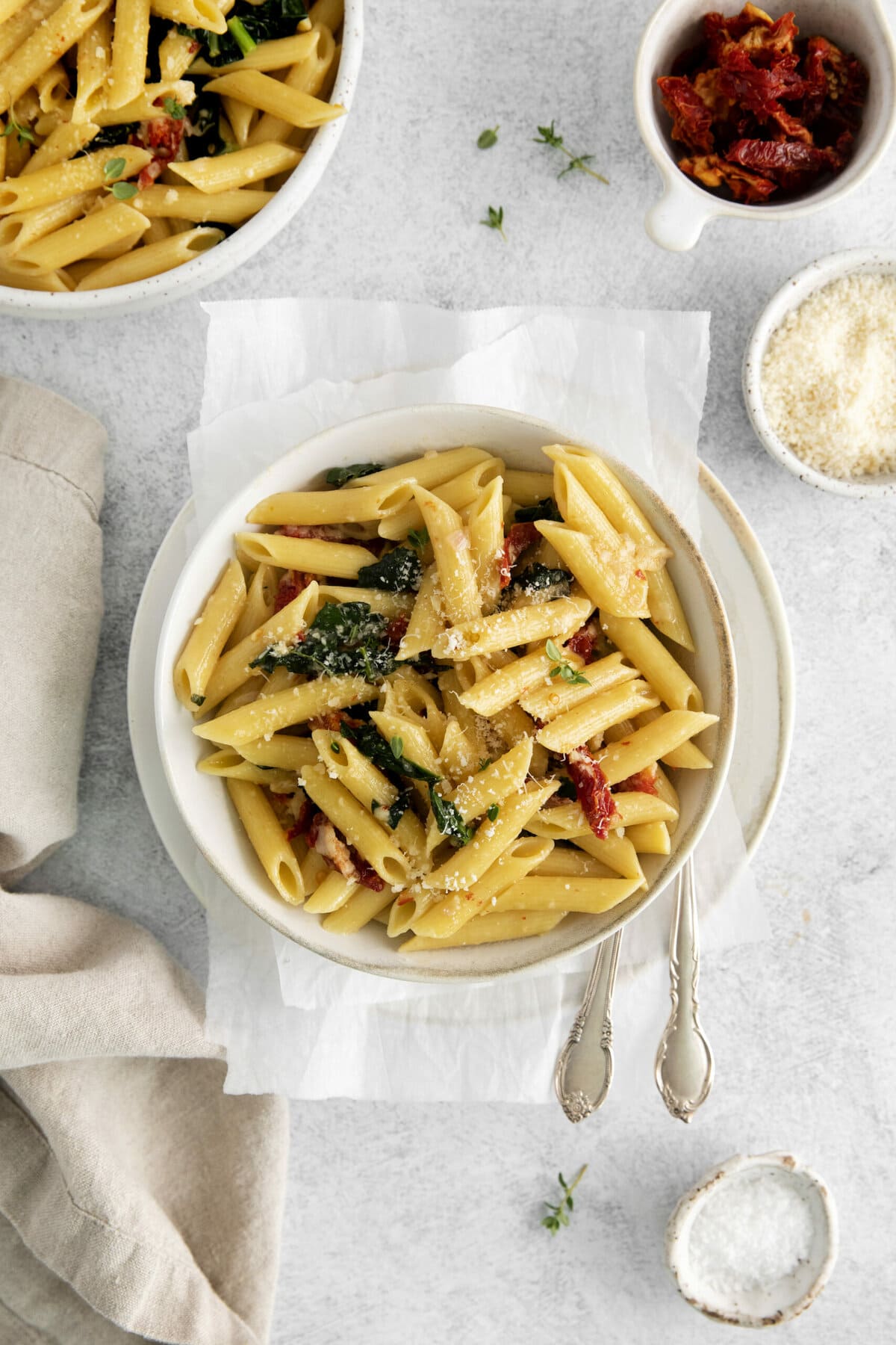 picture of penne pasta in a bowl with kale, sundried tomatoes, and cheese on a table with forks