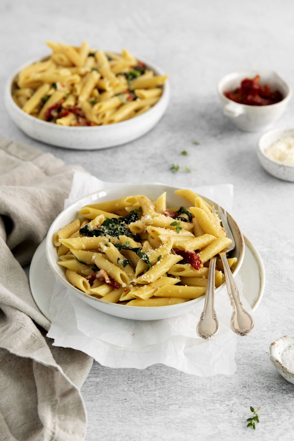 picture of penne pasta in a bowl with kale, sundried tomatoes, and cheese on a table with forks