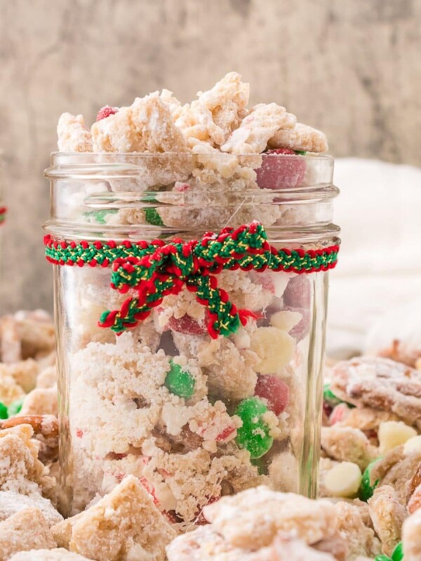 picture of muddy buddies chex mix with pretzels and red and green candies mixed in coated in powdered sugar in a jar with a red and green bow on it