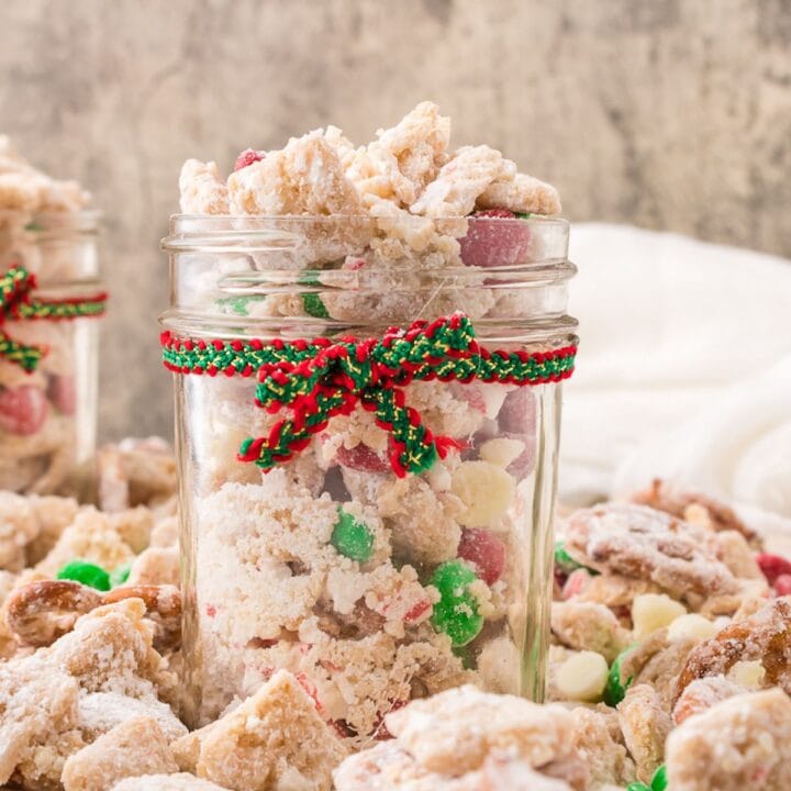 picture of muddy buddies chex mix with pretzels and red and green candies mixed in coated in powdered sugar in a jar with a red and green bow on it