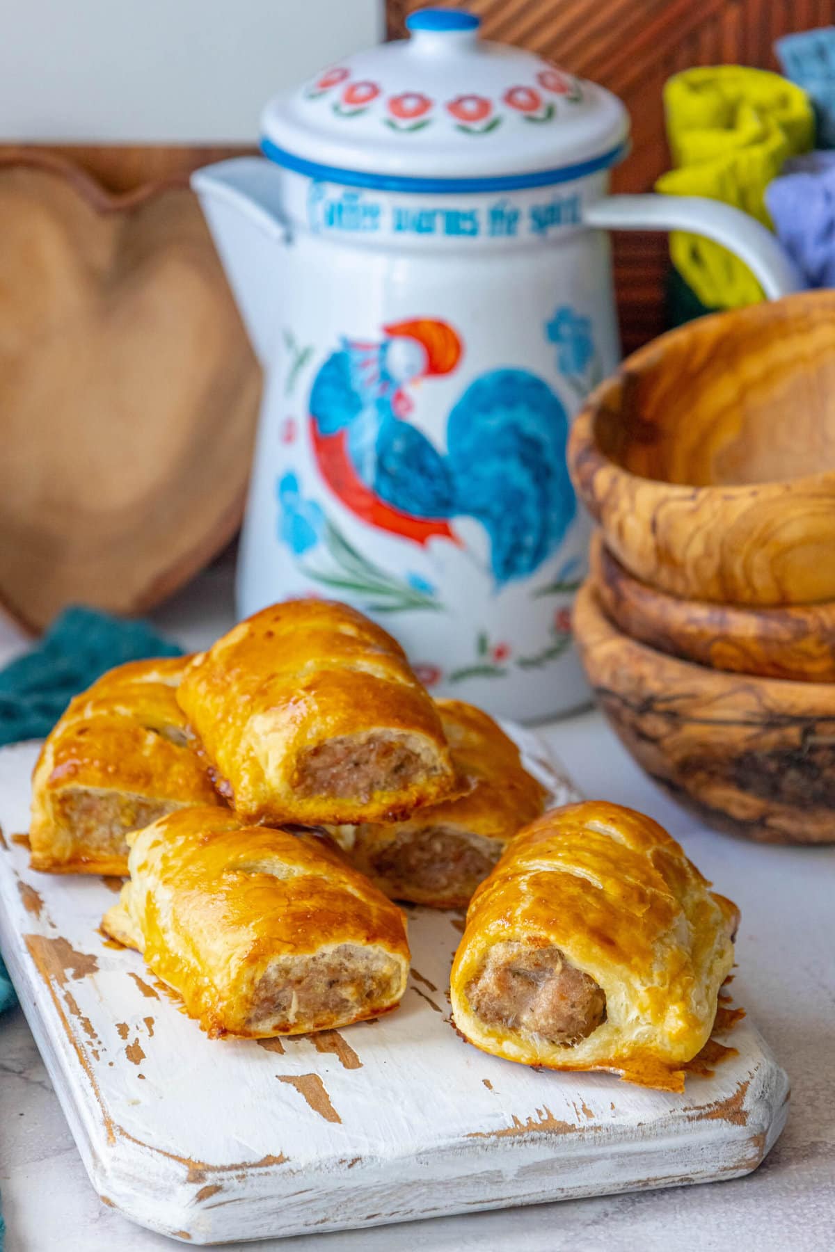 sausage rolls on a white cutting board with a rooster tea pot in the background