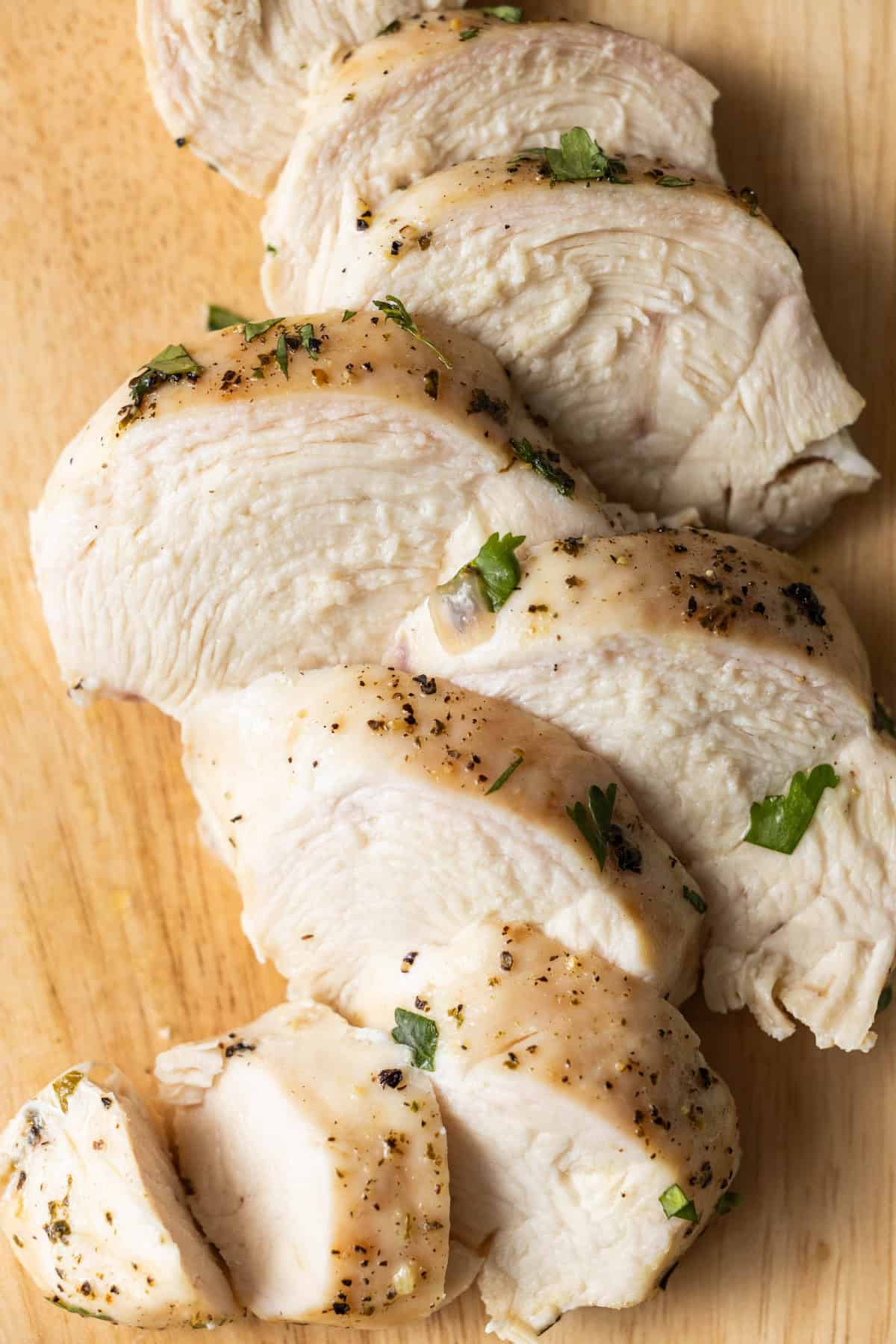 Sliced, easy baked chicken breast seasoned with herbs and black pepper on a wooden cutting board.