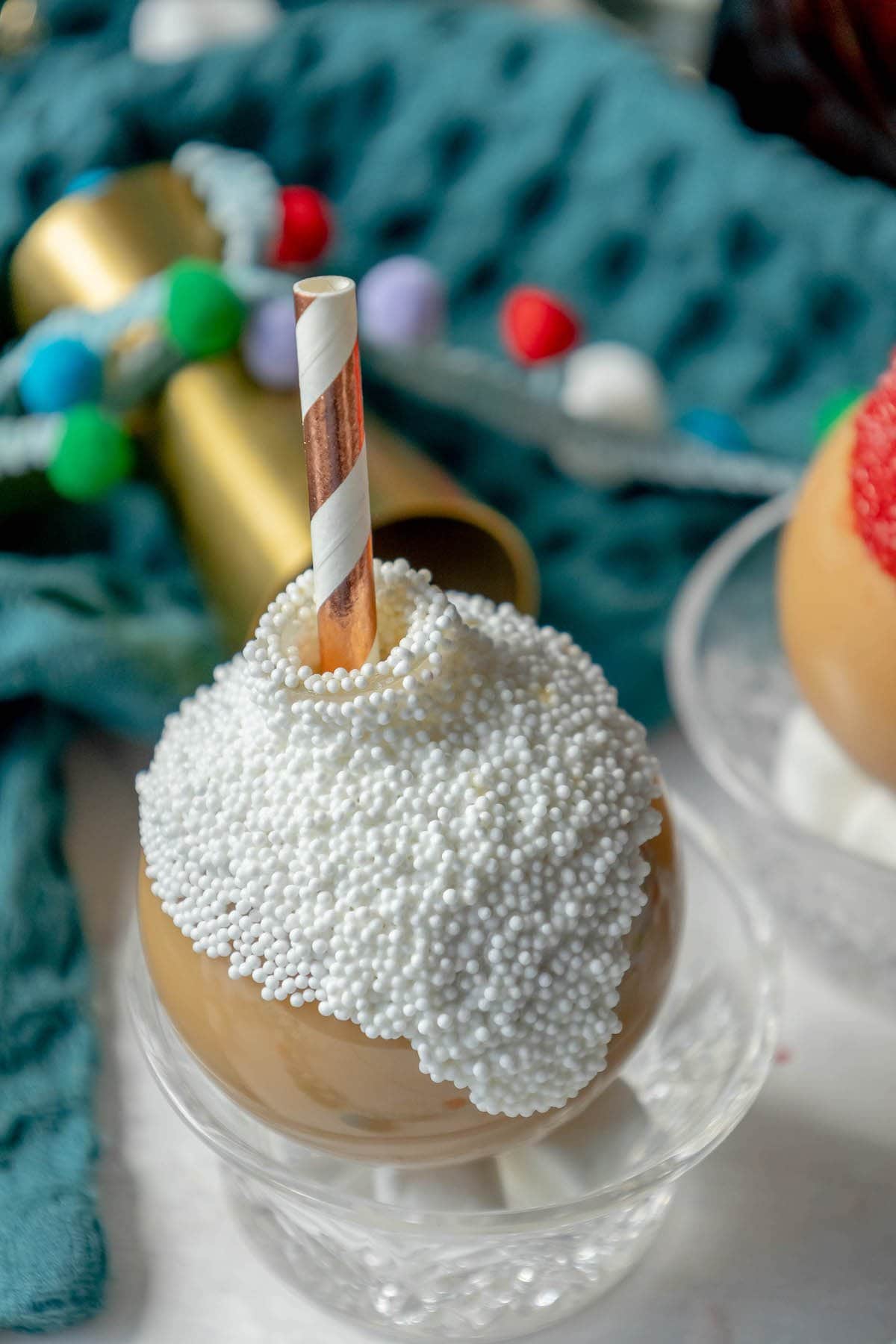 chocolate caramel whiskey cocktail in a plastic ornament with white chocolate and white nonpareil sprinkles and a straw inside in front of christmas decor