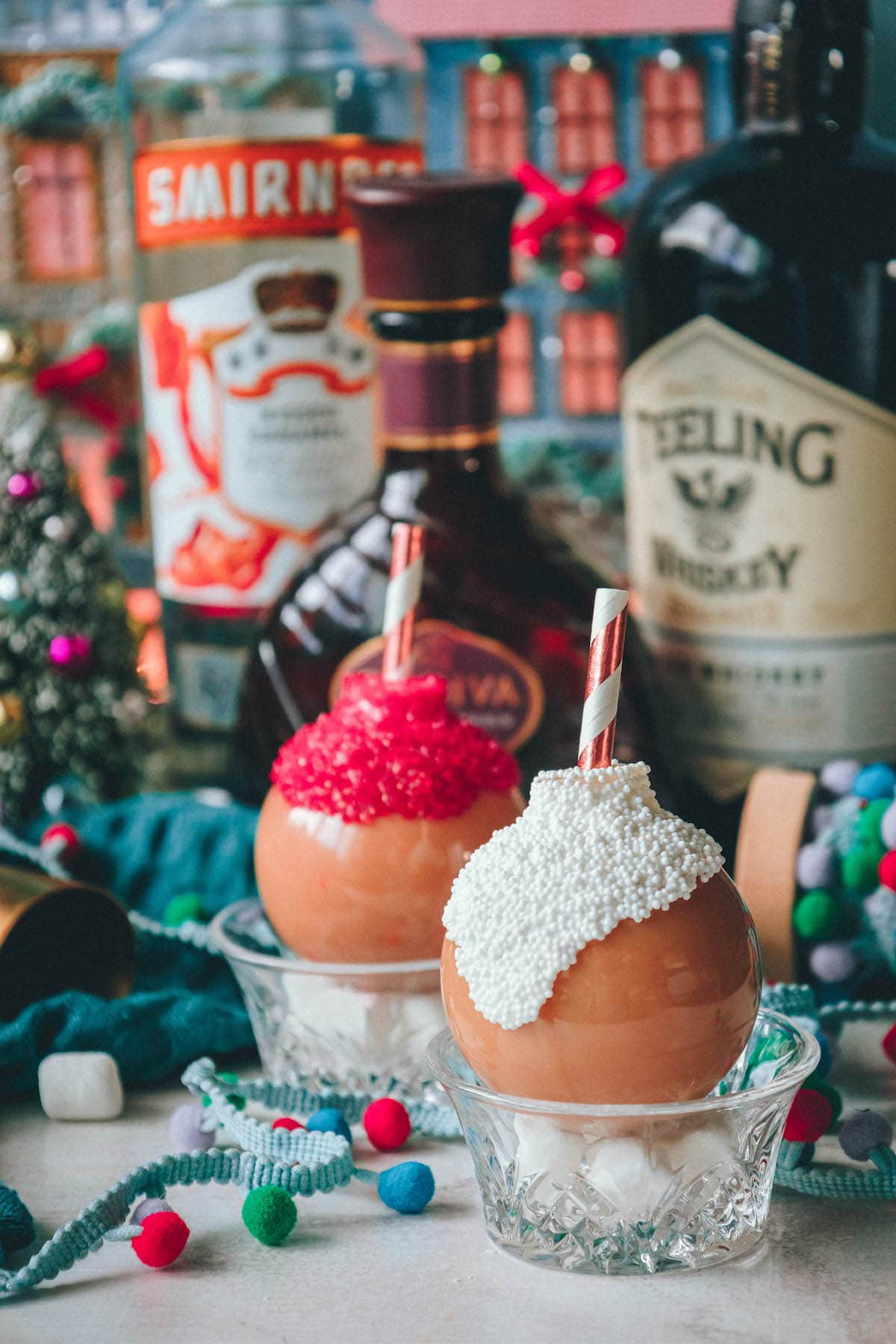 chocolate whiskey cocktails in plastic ornaments dipped in white chocolate covered with sprinkles with a straw inside on a table with Christmas decor