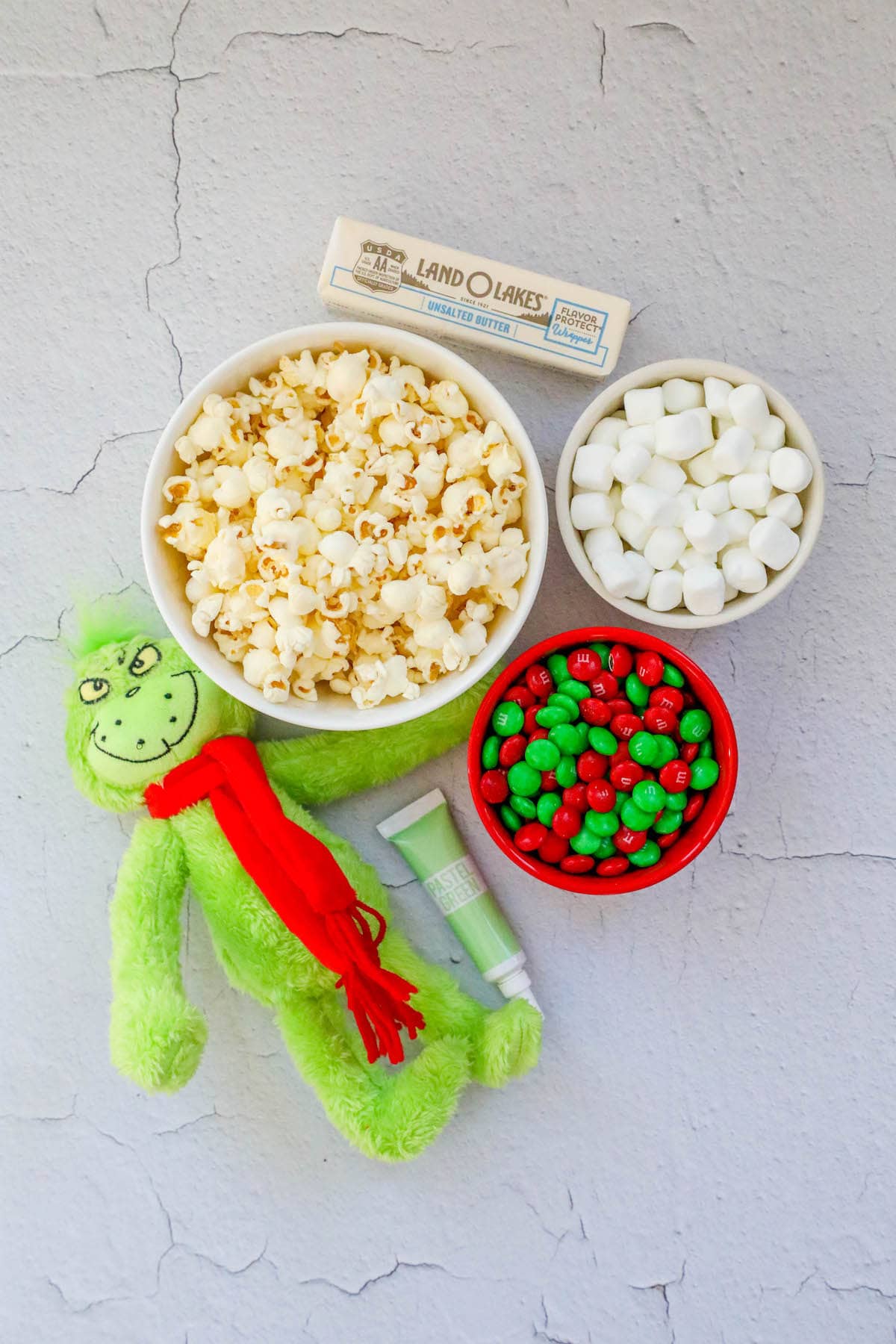 a grinch doll, bowl of popcorn, food coloring, m&m candies, marshmallows, and butter on a table