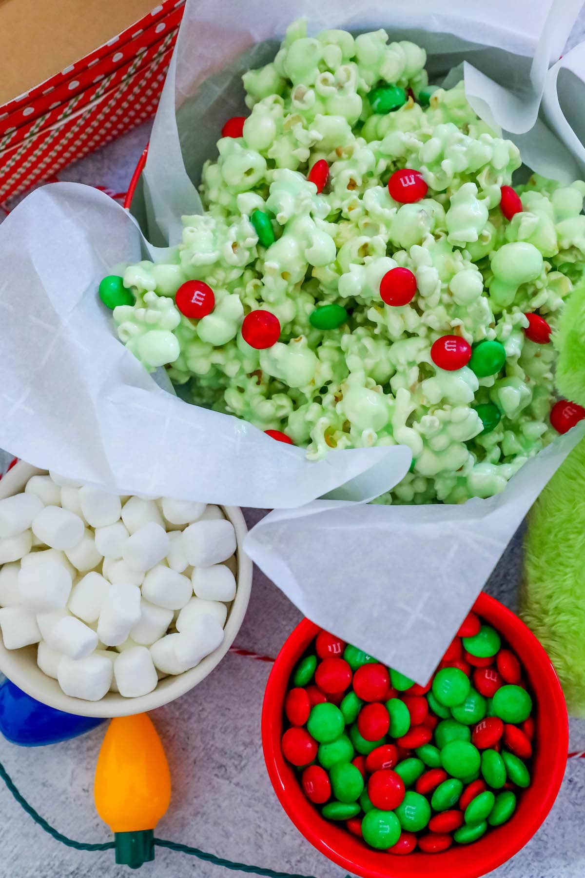 green candy coated popcorn with green and red christmas candies in a bowl on a table with marshmallows, christmas lights and m&ms