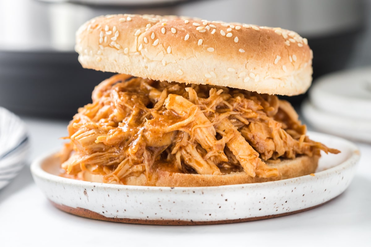 shredded chicken sandwich with bbq sauce in a sesame bun on a white plate in front of an instant pot