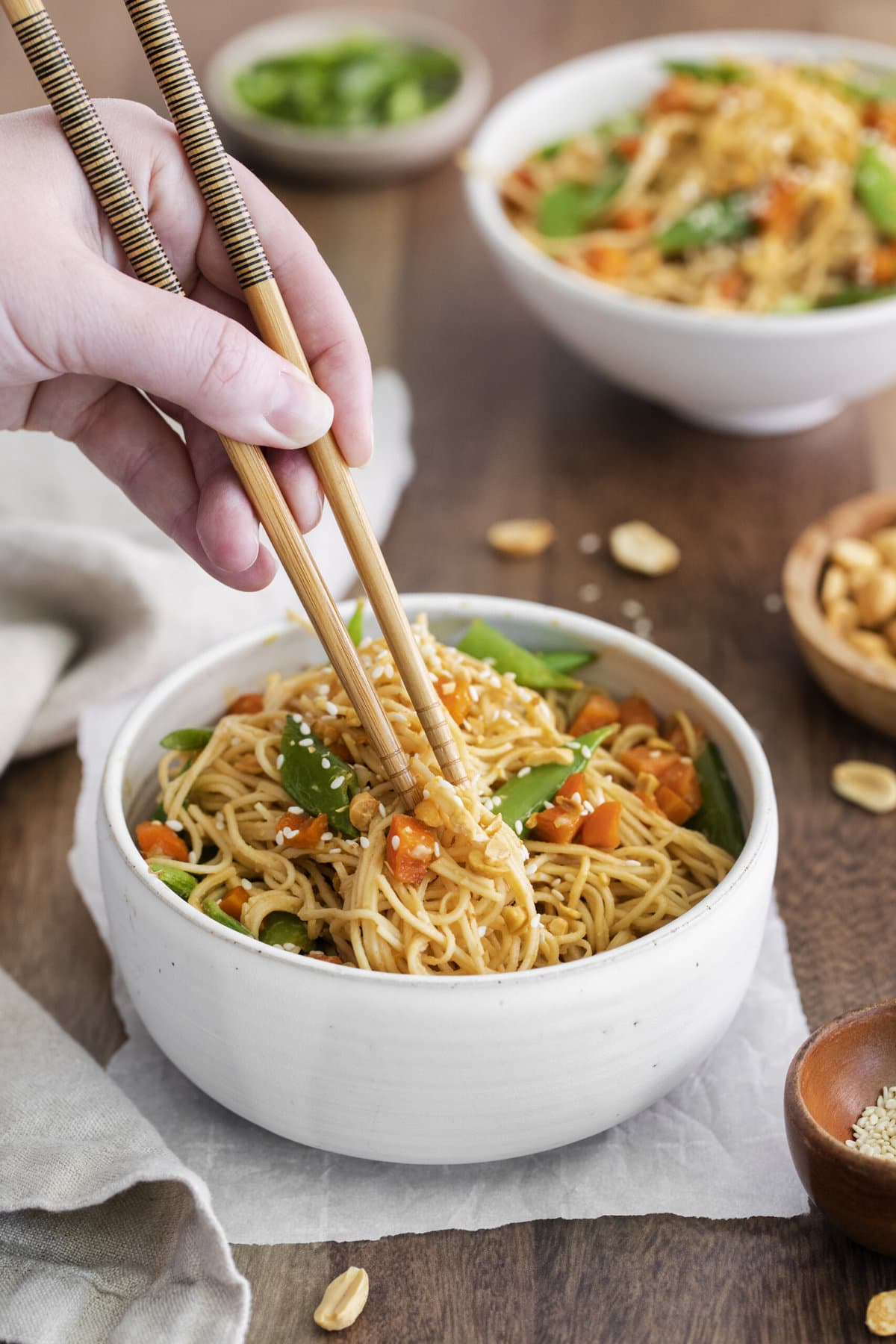 peanut noodles with peas and carrots in a white bowl, someone using chop sticks to take some out.
