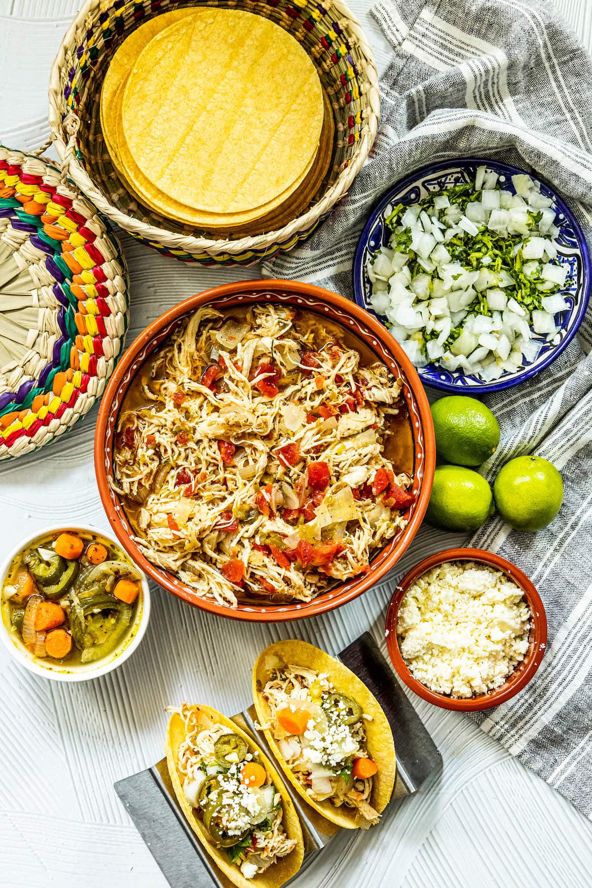 shredded chicken with tomatoes and green chile in a telavera bowl next to chicken tacos in a taco stand and a bowl of jalapeno and carrot salsa, a bowl of crumbled white cheese, some limes, a bowl of diced onions, and a bowl of tortillas on a table