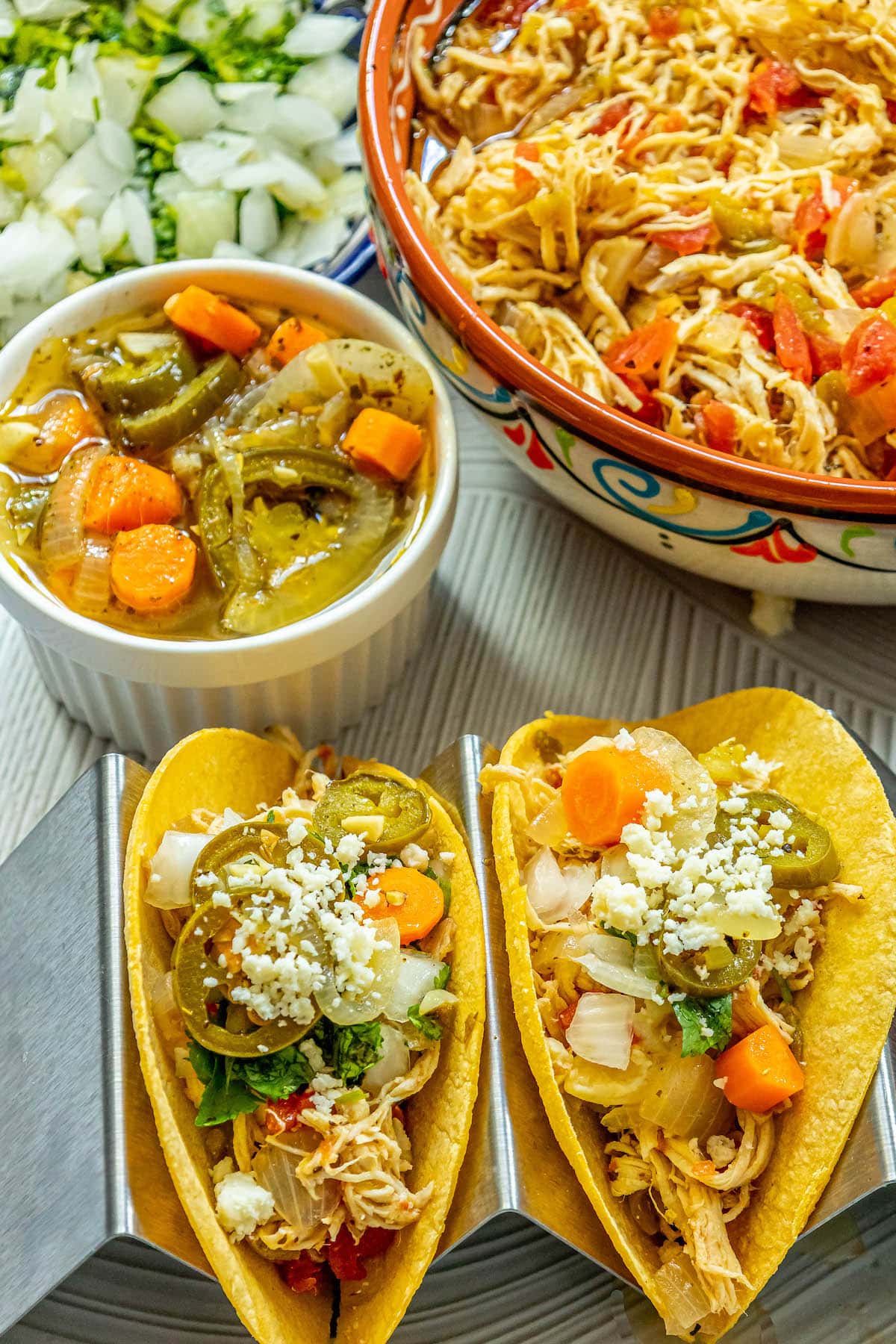 chicken tacos with shredded chicken, pickled jalapenos, crumbled white cheese, onions, chopped cilantro, diced onion, and pickled carrot in a metal stand in front of a bowl of pickled carrot and jalapeno salsa