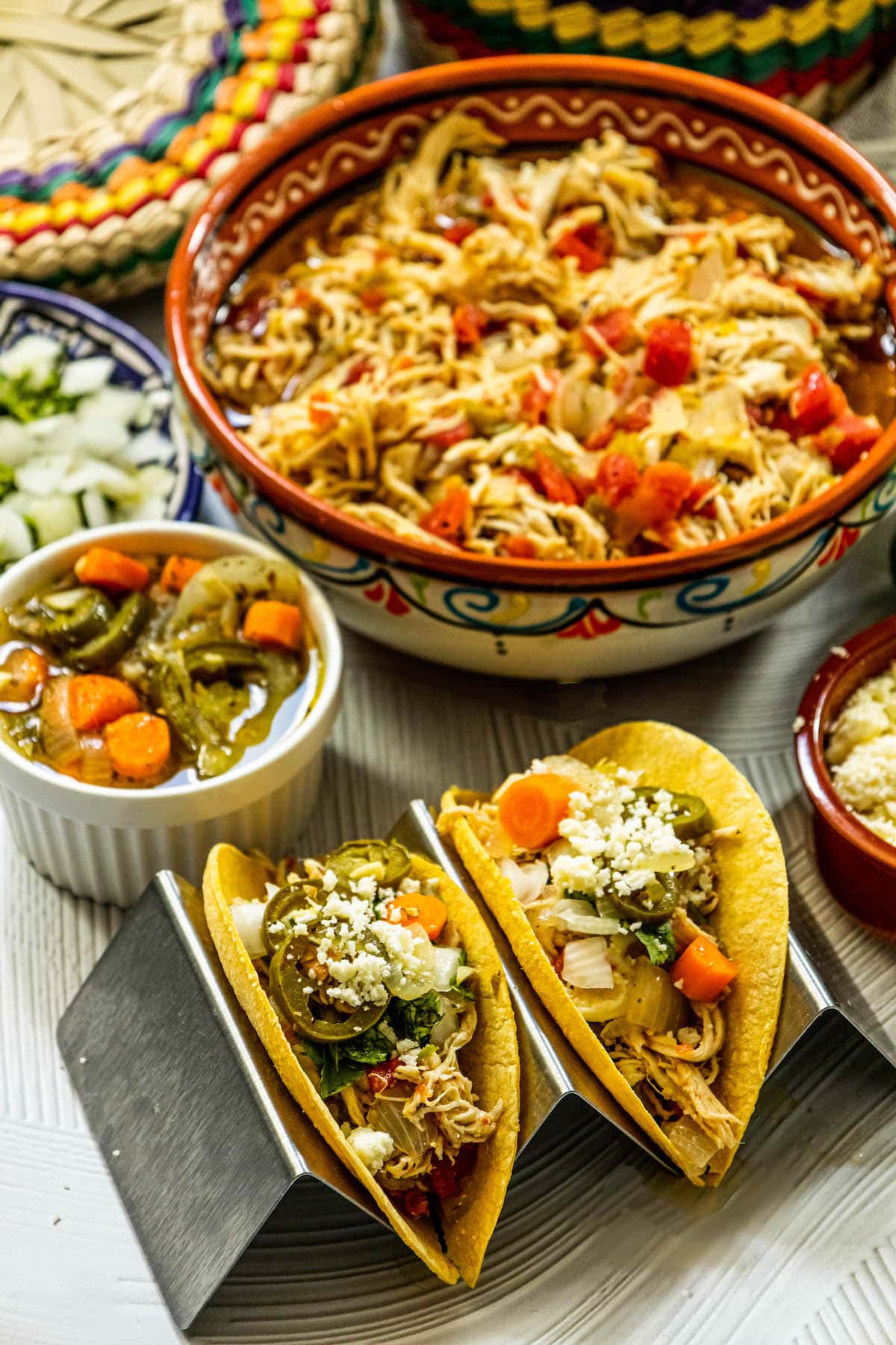 shredded chicken with tomatoes and green chile in a telavera bowl next to chicken tacos in a taco stand and a bowl of jalapeno and carrot salsa, a bowl of crumbled white cheese, some limes, a bowl of diced onions, and a bowl of tortillas on a table