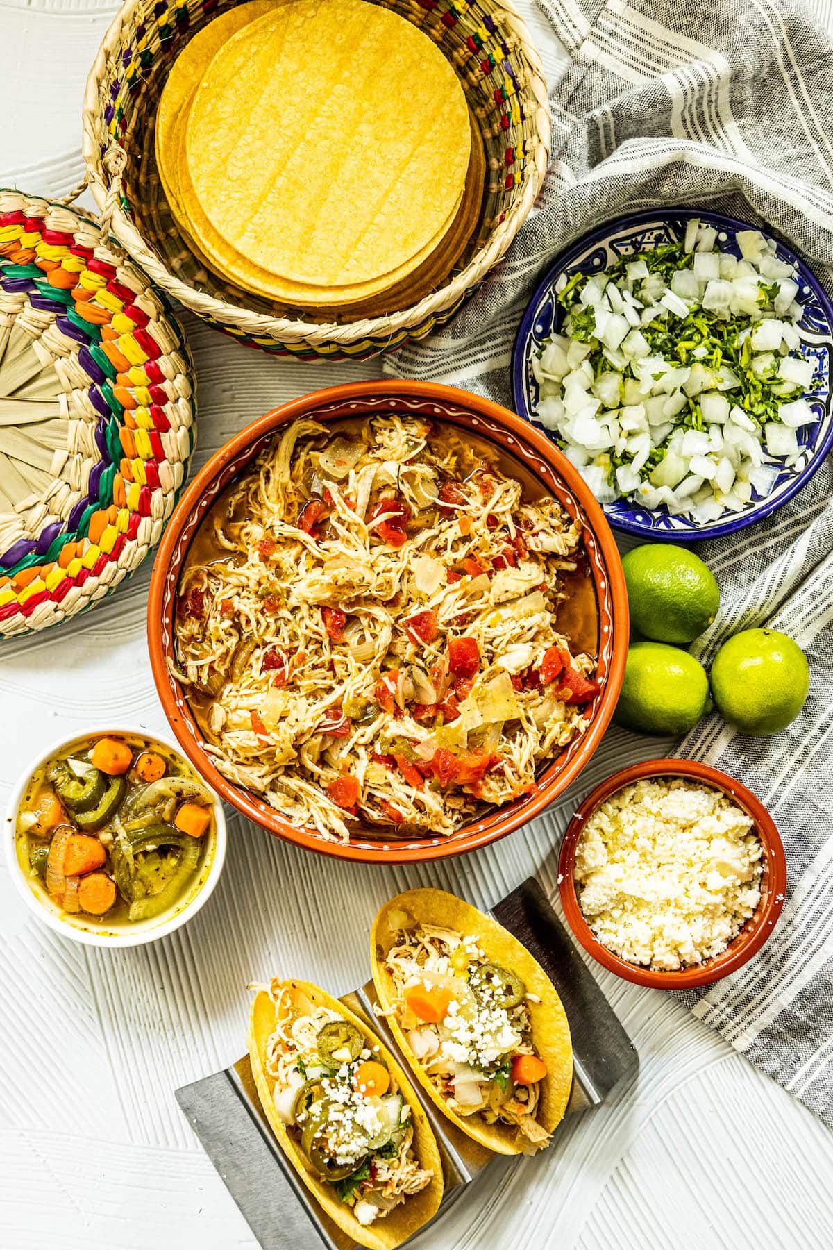 shredded chicken with tomatoes and green chile in a telavera bowl next to chicken tacos in a taco stand and a bowl of jalapeno and carrot salsa, a bowl of crumbled white cheese, some limes, a bowl of diced onions, and a bowl of tortillas on a table