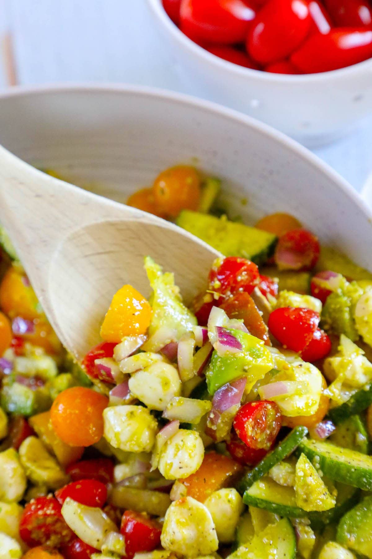 Diced cucumber, red onion, grape tomatoes, feta cheese, avocados, and basil in a pesto sauce in a white bowl 