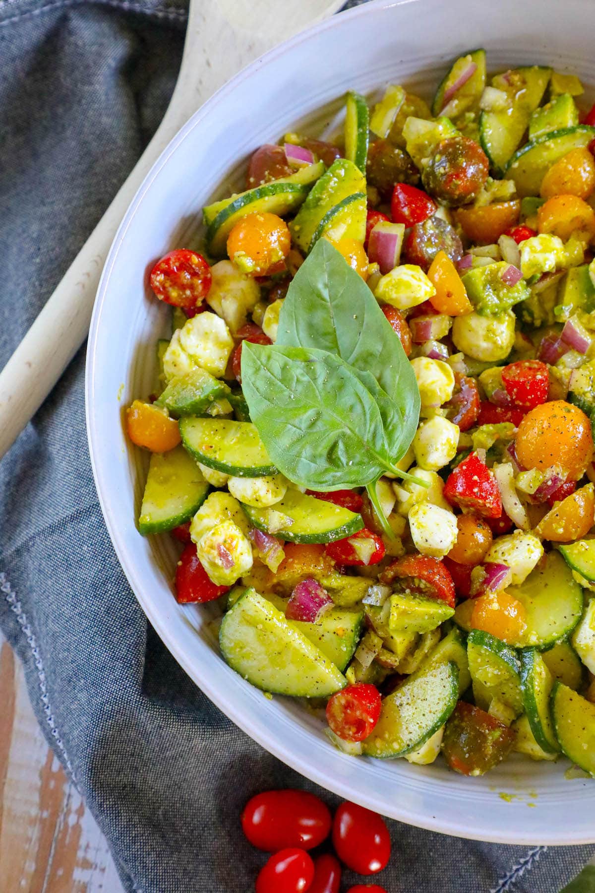 Diced cucumber, red onion, grape tomatoes, feta cheese, avocados, and basil in a pesto sauce in a white bowl 