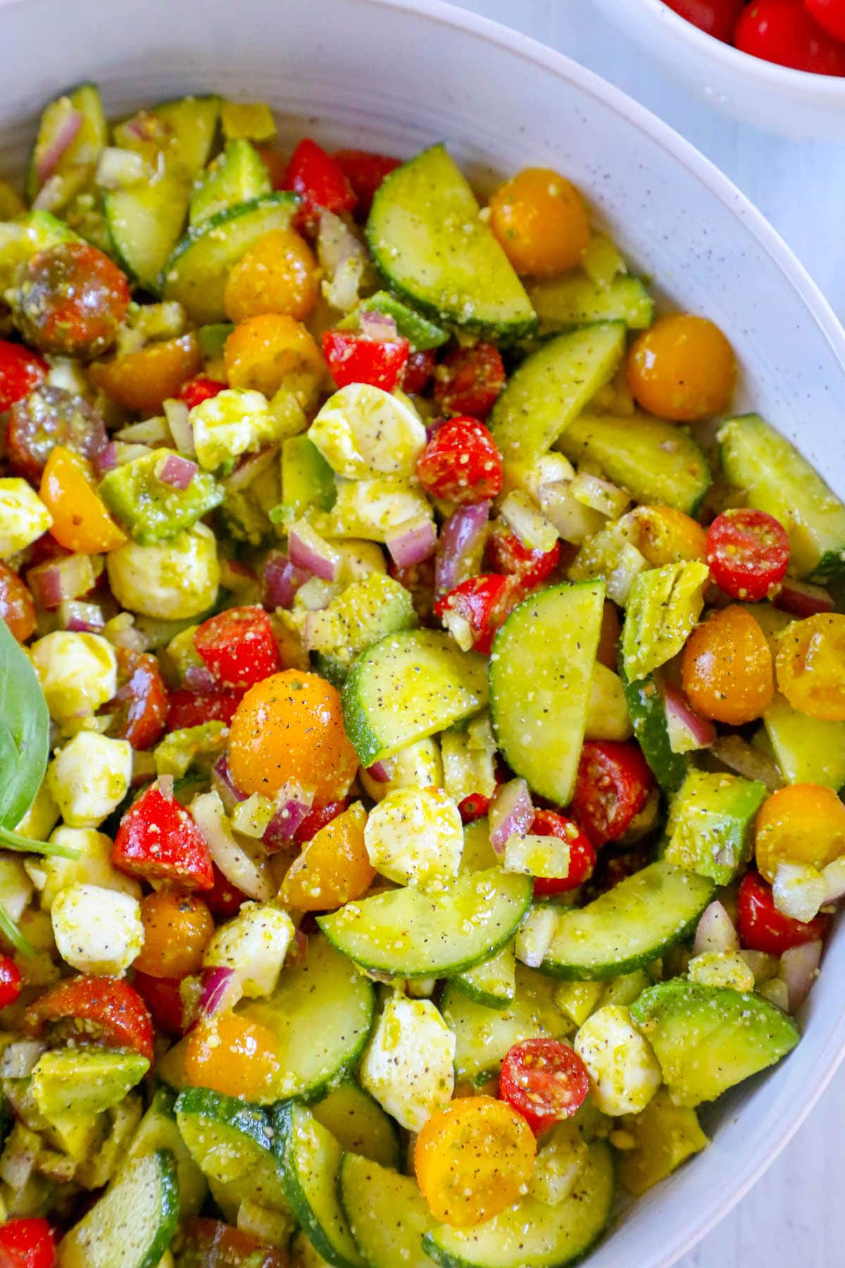 Diced cucumber, red onion, grape tomatoes, feta cheese, avocados, and basil in a pesto sauce in a white bowl 