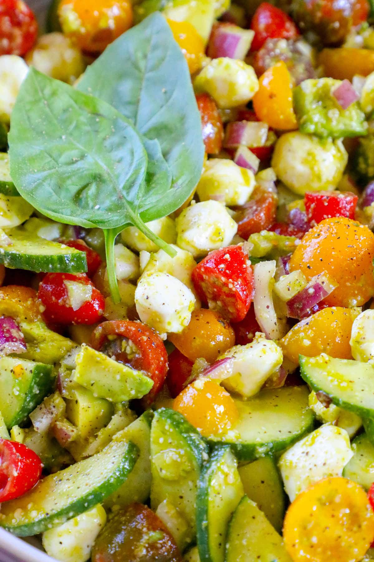 Diced cucumber, red onion, grape tomatoes, feta cheese, avocados, and basil in a pesto sauce in a white bowl 