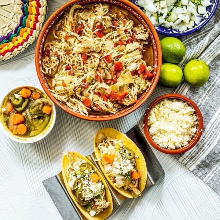 shredded chicken with tomatoes and green chile in a telavera bowl next to chicken tacos in a taco stand and a bowl of jalapeno and carrot salsa, a bowl of crumbled white cheese, some limes, a bowl of diced onions, and a bowl of tortillas on a table