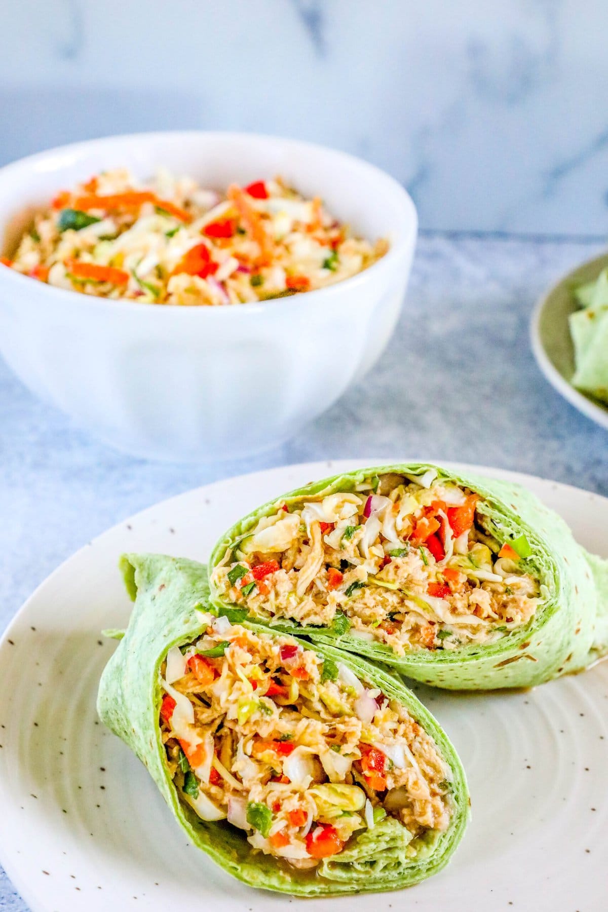 chicken salad with shredded chicken, cabbage, and diced vegetables wrapped in a green tortilla sliced in half on a plate in front of a bowl of chicken salad