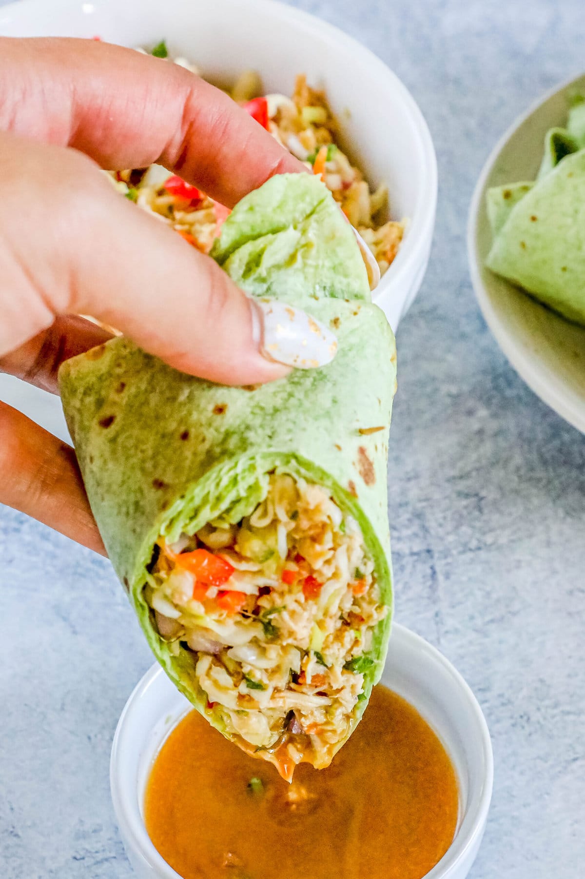 a hand dipping a Thai chicken salad wrap in a green spinach tortilla in a small cup of peanut sauce