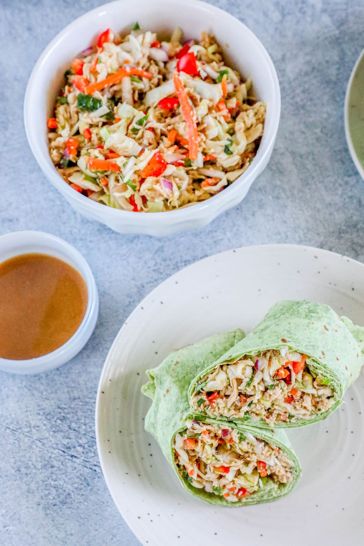 chicken salad with shredded chicken, cabbage, and diced vegetables wrapped in a green tortilla sliced in half on a plate next to a cup of peanut sauce