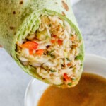 chicken salad with shredded chicken, cabbage, and diced vegetables wrapped in a green tortilla sliced in half being dipped into a bowl of peanut sauce