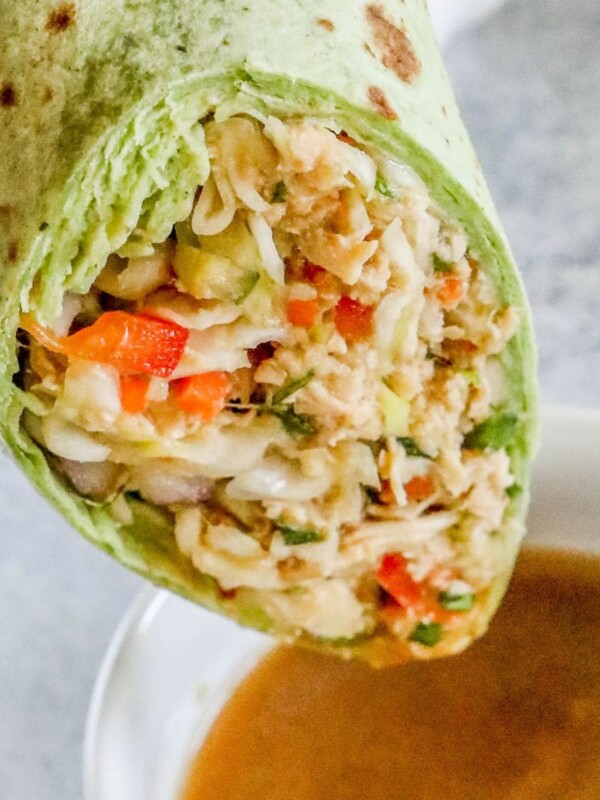 chicken salad with shredded chicken, cabbage, and diced vegetables wrapped in a green tortilla sliced in half being dipped into a bowl of peanut sauce