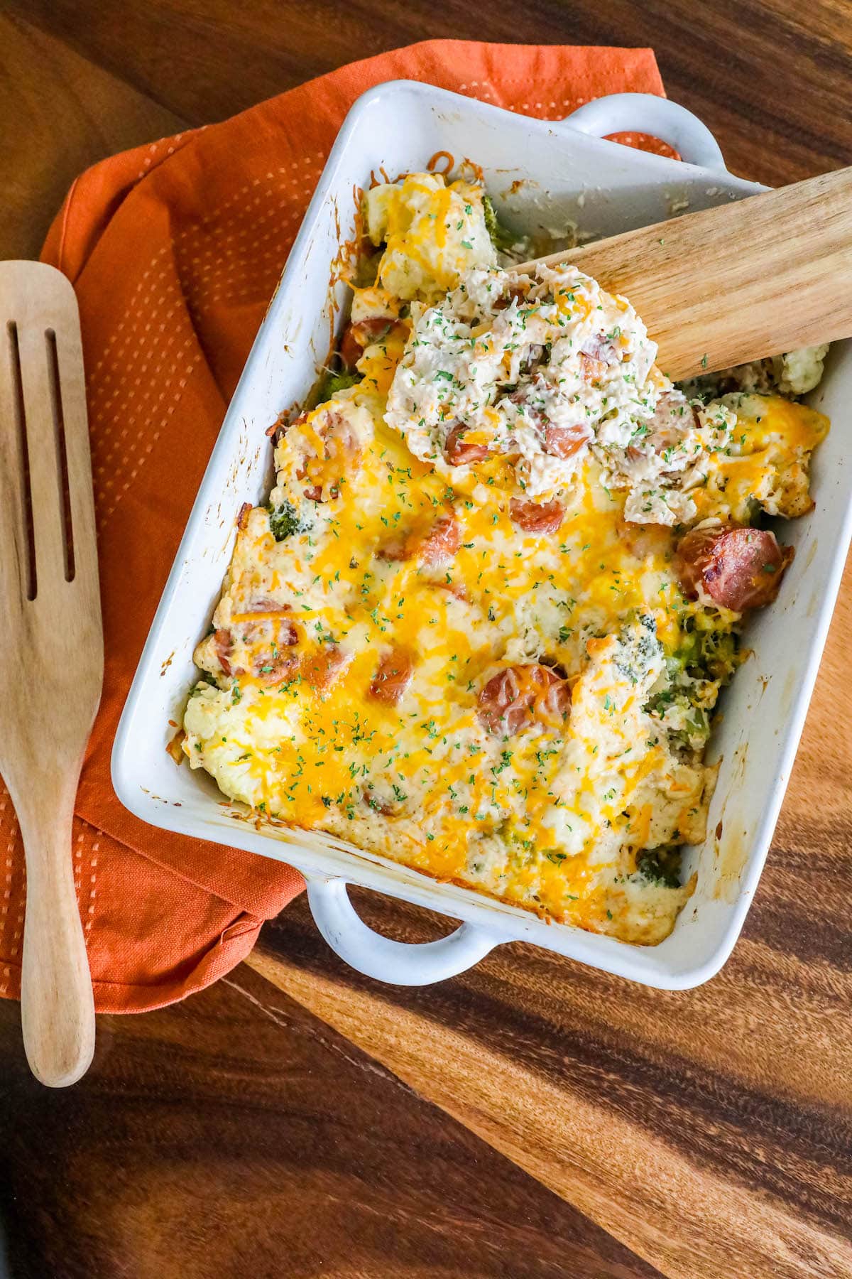 a spoon scooping up cheesy casserole with sliced sausage, broccoli, cauliflower, and herb seasonings in a white casserole dish on a table
